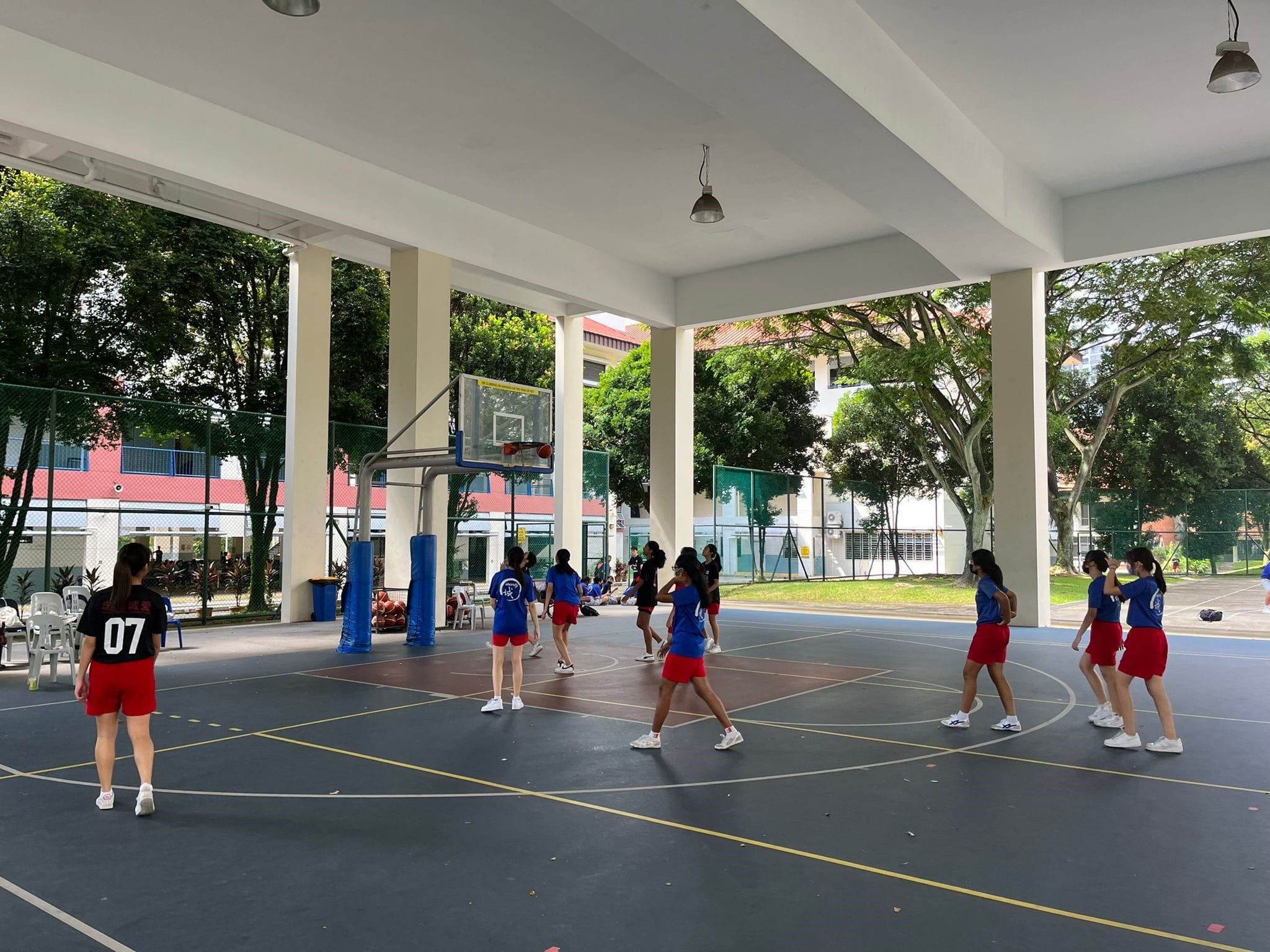 Girls in blue shirts and red shorts play on a covered outdoor basketball court near school buildings.